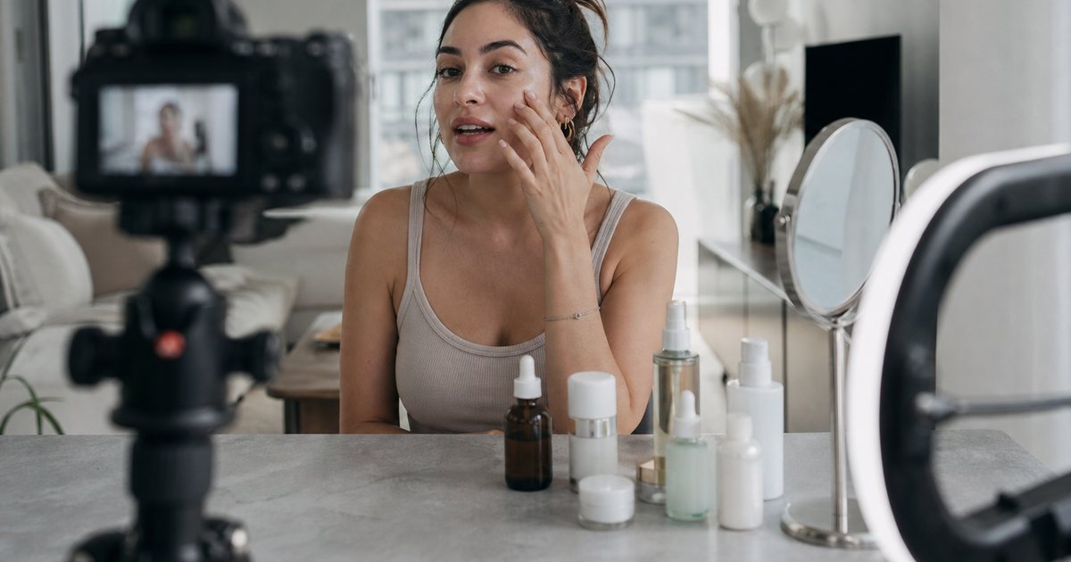 A young woman filming herself with a camera on a tripod in a bright apartment, skincare bottles and a ring light on the table, seen from behind the camera