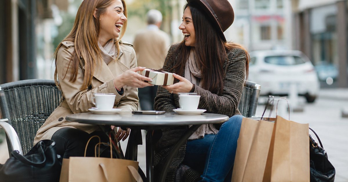 Two friends laughing at an outdoor cafe table, paper shopping bags at their feet, one handing the other a small wrapped gift box, city street in soft focus