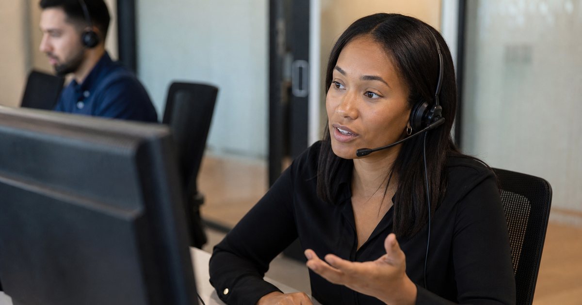 A customer support agent wearing a headset and speaking, seated at a desk in a small office with a notebook and coffee mug, monitor turned away from the camera, colleague in the background